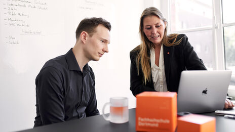 Foto von zwei Personen, die zusammen an einem Tisch sind und gucken gemeinsam auf den Bildschirm eines Notebooks. Eine Person sitzt am Tisch und die andere steht am Tisch. Die am Tisch stehende Person erzählt der anderen Person etwas. Auf dem Tisch stehen zwei orangene FH Würfel und eine FH Tasse.