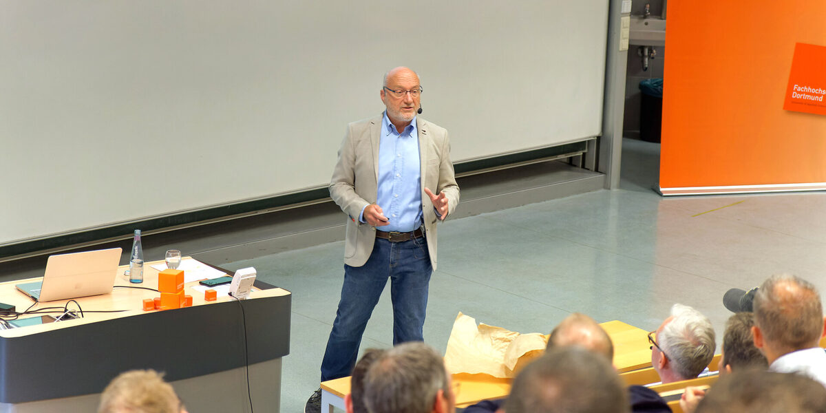 A man stands in a lecture hall and speaks to the people sitting in front of him.