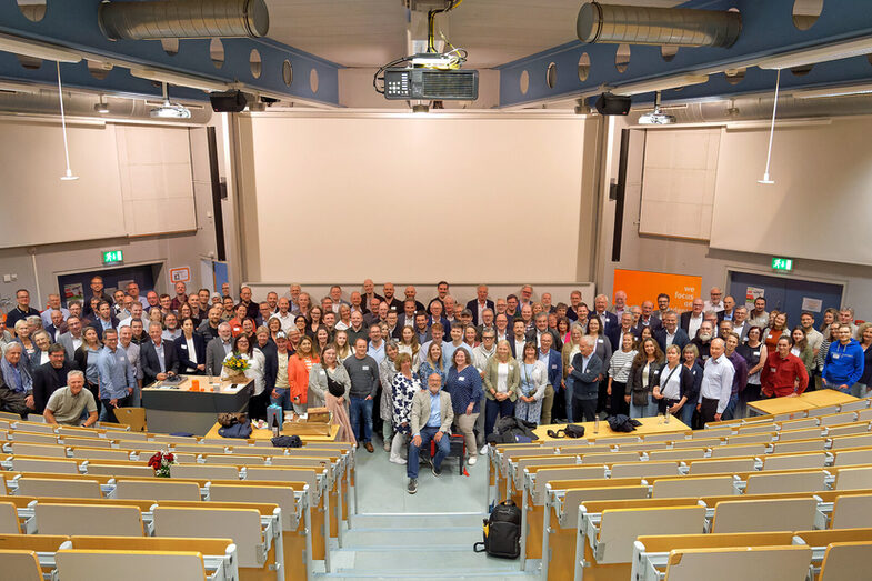 A large group of people stand together for a group photo in the lecture hall of the Faculty of Computer Science. One person is sitting on a piano stool at the front.