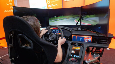 One person sits in the driving simulator, controls the steering wheel and looks at three screens.