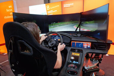 One person sits in the driving simulator, controls the steering wheel and looks at three screens.