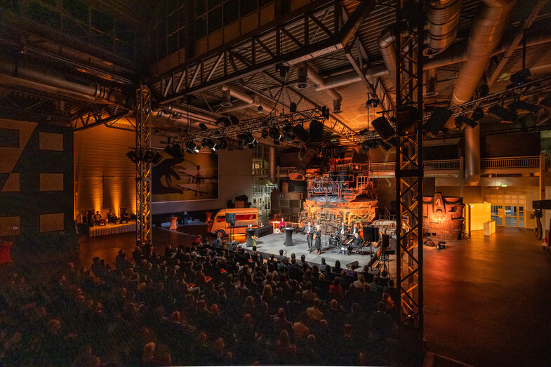 Foto einer illuminierten Halle. In dieser ist eine Bühne aufgebaut, davor sitzt Publikum.