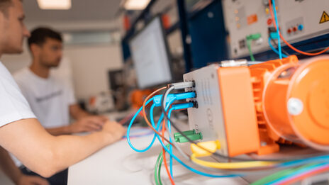 Foto von einem Elektromotor, im Hintergrund arbeiten 2 Studierenden am Computer des Prüfstandes.