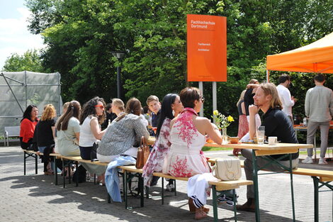 View of the table set up in front of the building, where former students are chatting animatedly, with the orange UAS pavilion in the background.