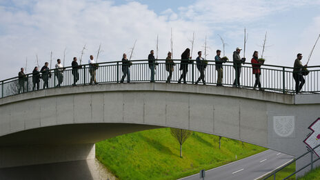 Several people walk one after the other across a pedestrian bridge, each holding a small plant.