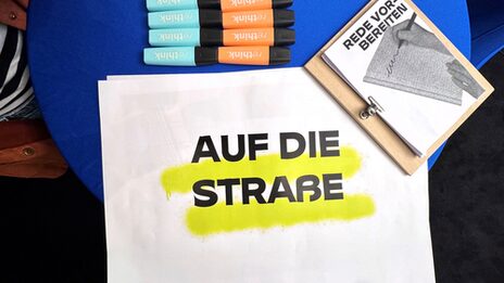 Auf einem Tisch liegt ein Plakatentwurf mit der Aufschrift "Auf die Straße".