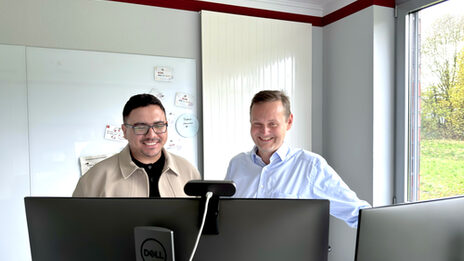 Zwei Personen stehen hinter zwei Monitoren und blicken darauf.