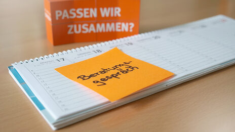 Foto eines Tischkalenders, auf dem ein orangefarbenes Post-It mit der Notiz „Beratungsgespräch“ klebt. Im Hintergrund stehen gestapelte Würfel, auf denen „Passen wir zusammen?“ steht.