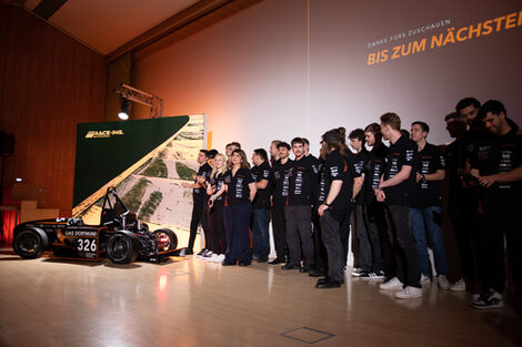 Around 20 people in black T-shirts are standing on a stage, with a racing car to their left.