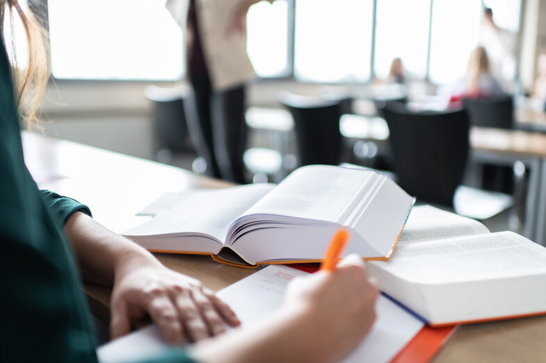 Photo of a woman taking notes. There are two books in front of her. In the background are several people in the seminar room. __ Woman takes notes. There are two books in front of her. In the background there are several people in the seminar room.