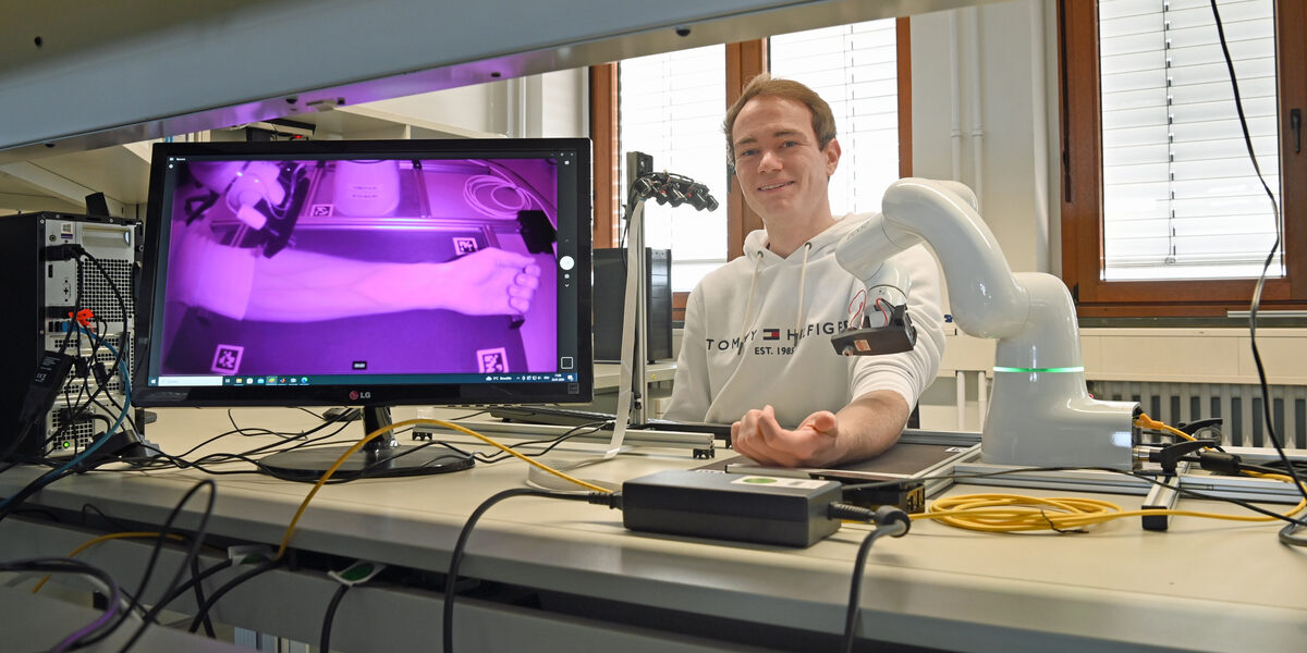 A person is sitting at a laboratory table and has placed their forearm under a robot. The veins of the forearm are clearly visible in infrared light on a screen next to it.