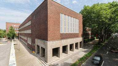 Foto mit Blick auf Teile eines Gebäudes der Fachhochschule Dortmund am Max-Ophüls-Platz mit Vorplatz und Gehweg sowie Straße mit Autos daneben.