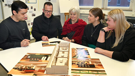 A group of people sits around a model and illustrations and discusses the designs.