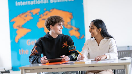Photo of two students sitting at a table and talking. In front of them on the table are work materials. On the wall behind them hangs an orange world map on a blue background with the heading "International Business".