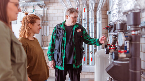 Photo of 3 people standing in front of a heating system. The man seems to be explaining something to the two women.