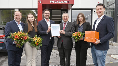 Six people stand next to each other, looking friendly into the camera, in the background a short flight of stairs leading to the entrance of a store with the word "Weckbacher" on the canopy. Three people hold a bouquet of flowers in their hands, the two in the middle hold a certificate up to the camera.