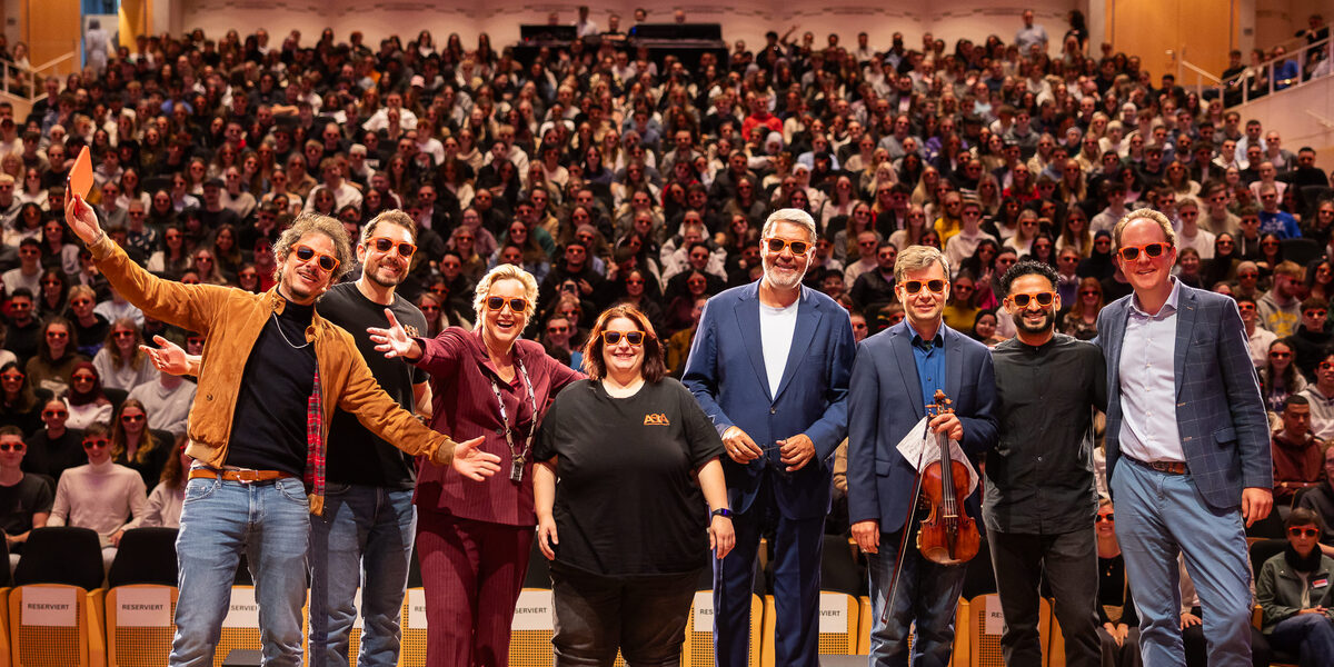 Mehre Personen mit Sonnenbrille stehen auf einer Bühne. Im Hintergrund ist ein voller Saal zu sehen.