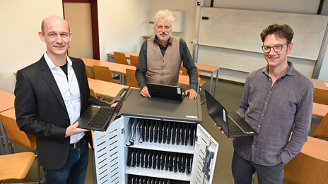 Three people are standing around a roll container filled with notebooks. Two of them are holding a laptop.