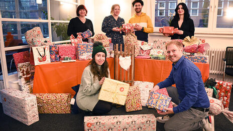 Four people are standing behind a table covered in orange with lots of wrapped presents, each holding presents in their hands. Two more people are sitting in front of the table. They are all smiling and looking at the camera.