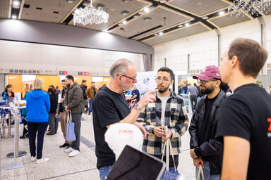 Mehrere Personen im Gespräch miteinander. Im Hintergrund sind weitere Messestände zu sehen.