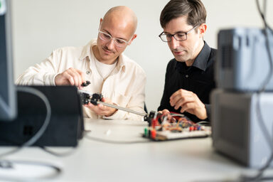 Bild von einem Studierenden neben seinem Professor. Beide sitzen an einem Versuchsaufbau.