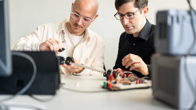 Picture of a student next to his professor. Both are sitting at an experimental setup.