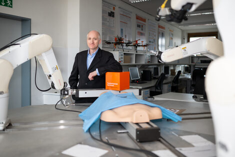 Foto von einem Mann, der hinter einem Aufbau von einem OP-Roboter steht und mit dem linken Arm auf einem orangenen Würfel lehnt.