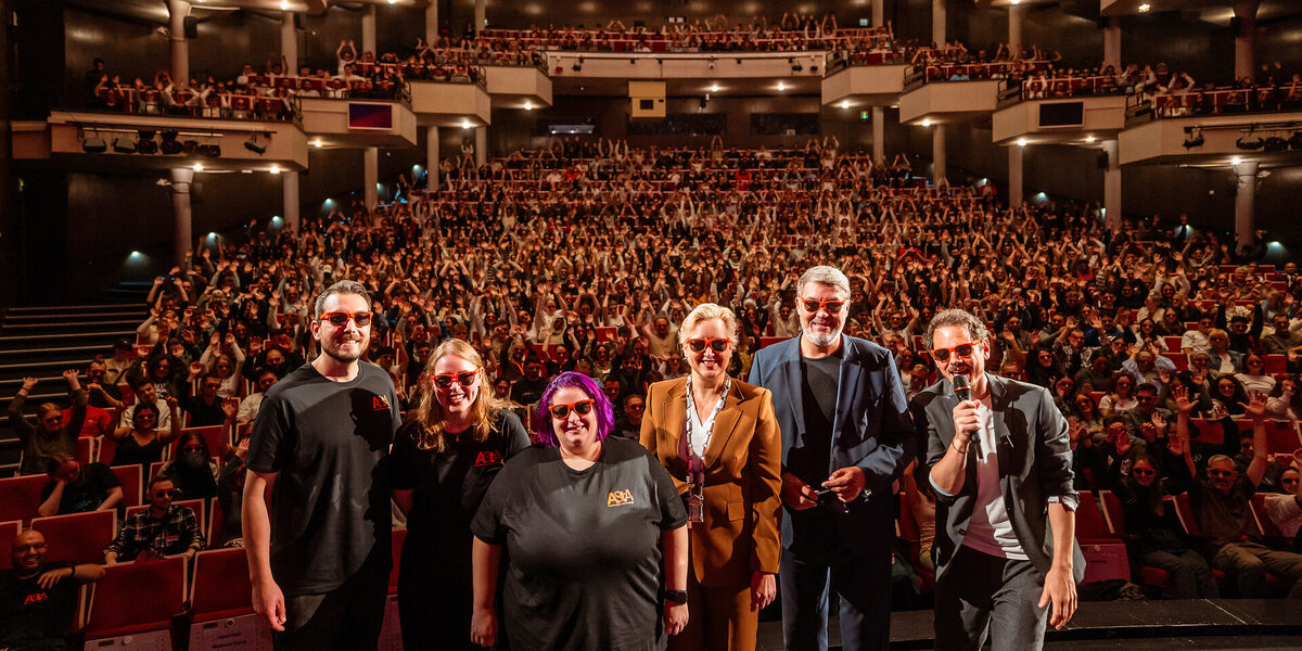 Several people wearing sunglasses on stage in the opera house. In the background, the first-semester students in the stands make "la Ola".