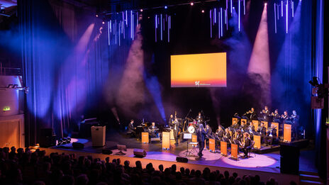 A big band plays on the stage in the concert hall. The music stands are wrapped with "50 years of Fachhochschule Dortmund". The stage shines in orange light.