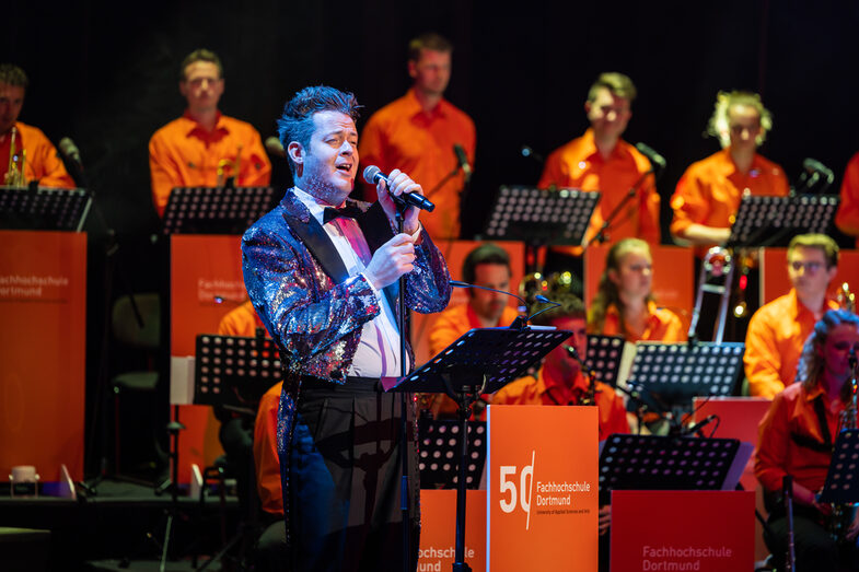 A singer with a glittering jacket sings. The band in orange shirts can be seen in the background.