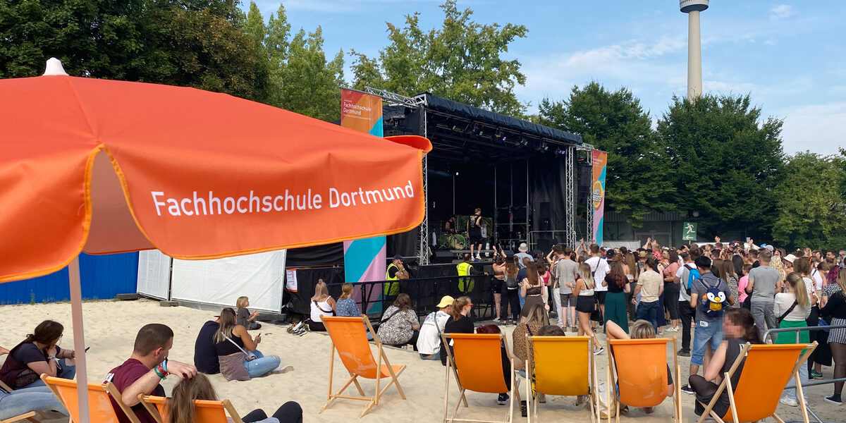 People sit in the FH Dortmund loungers under the parasols in front of a stage.