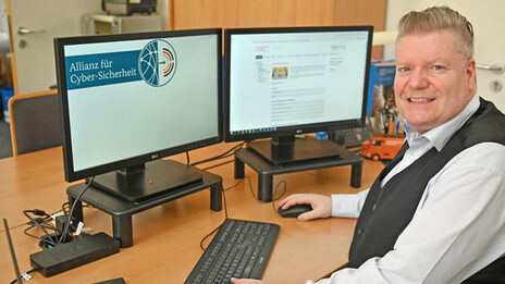 A male person is sitting at a desk in front of two monitors. The logo of the Alliance for Cyber Security can be seen on one monitor.