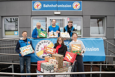 Sechs Personen stehen auf dem Aufgang zum Eingang der Bahnhofsmission Dortmund. Jede von ihnen hält ein festlich verpacktes Päckchen in den Händen. Vorn in der Mitte steht ein Einkaufswagen voll mit weiteren Geschenkpäckchen.