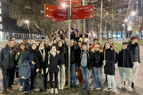 Rund 35 Menschen stehen bei abendlicher Dunkelheit auf einer hell beleuchteten Mittelinsel einer großstädtischen Straße an einem Wegweiser, der in Richtung mehrerer Städte weist. Auf dem obersten Schild steht "Partnerschaften der Stadt Dortmund".