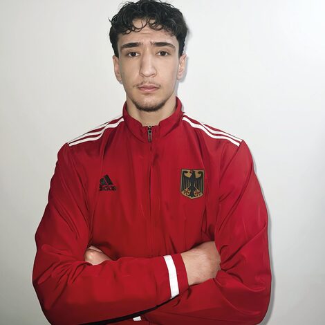 Ein Mensch in roter Trainingsjacke mit dem deutschen Wappen auf der Brust schaut mit verschränkten Armen in die Kamera.