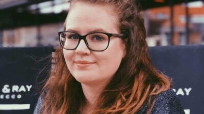 Gastwissenschaftlerin Dr. Charlotte Rottiers am Fachbereich Architektur. Sie trägt einen dunklen Pullover und lächelt leicht in die Kamera. Im Hintergrund sind unscharfe Schilder und städtische Strukturen zu sehen.