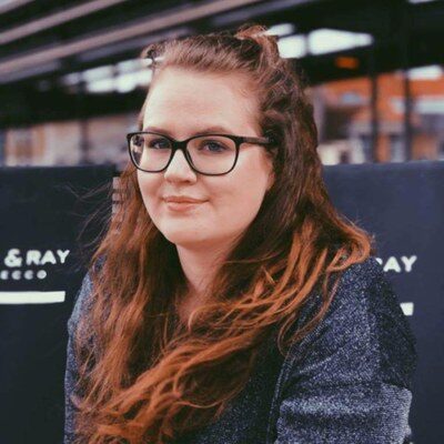 Visiting researcher Dr. Charlotte Rottiers at the Faculty of Architecture. She is wearing a dark sweater and smiling slightly at the camera. Blurred signs and urban structures can be seen in the background.