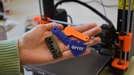 Four differently designed key rings lie in the palm of one hand, with a 3D printer in the background.