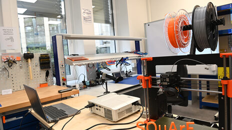 The picture shows a 3D printer with the printed three-dimensional SQuArE train; a workbench can be seen in the background.