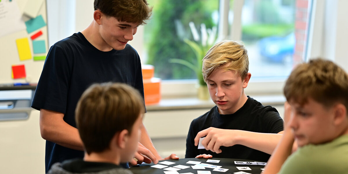 Four students at a high table with small cards representing data. One student holds up a card while the others watch.