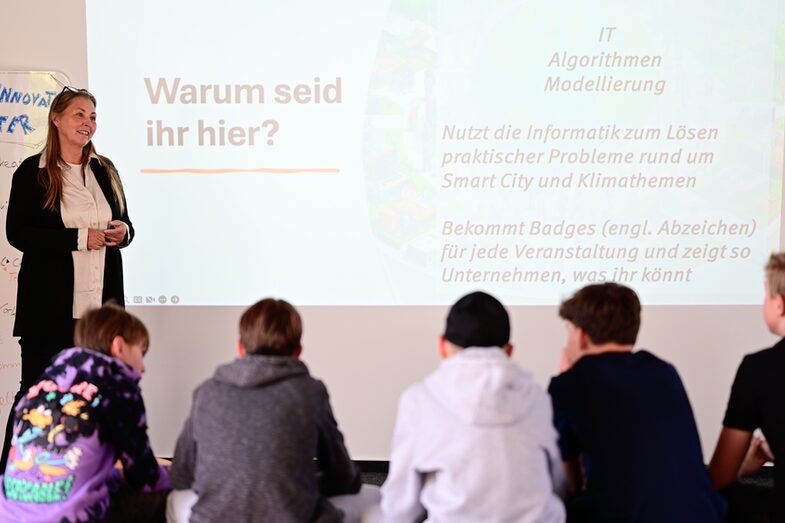 Prof. Dr. Sabine Sachweh gives an introductory lecture. She is standing in front of five students who are looking in her direction. On the wall behind her is a project with a slide. The caption reads: Why are you here?