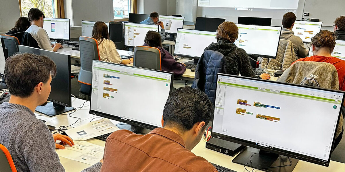 Mehrere Personen sitzen in einem Raum, vor ihnen stehen Computerbildschirme auf den Tischen.