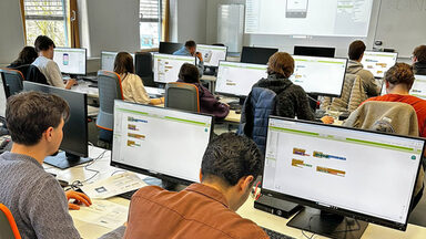 Several people are sitting in a room with computer screens on the tables in front of them.