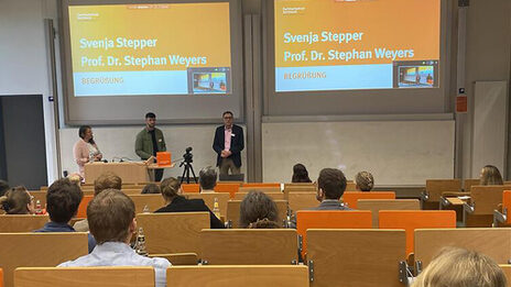 In a lecture hall, several seated people are looking at three people standing in front under two projector screens.