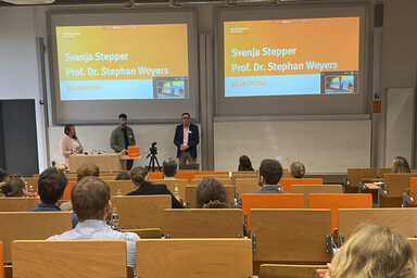 In a lecture hall, several seated people are looking at three people standing in front under two projector screens.
