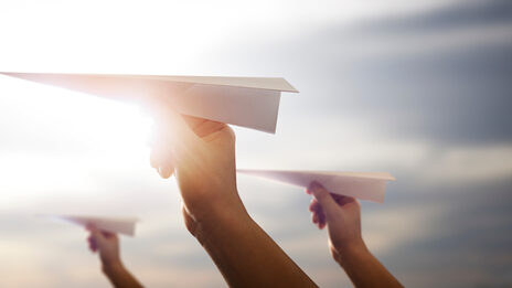 Foto von drei Händen die jeweils einen Papierflieger halten, vor einem sonnigen Himmel.