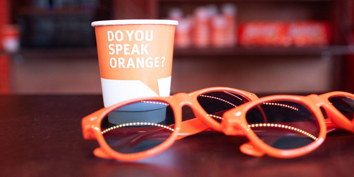 Auf einer Theke liegen zwei orangefarbene Sonnenbrillen. Daneben steht ein Becher mit der Aufschrift: Do you speak orange? Im Hintergrund sind verschwommen orangefarbene Objekte zu sehen.