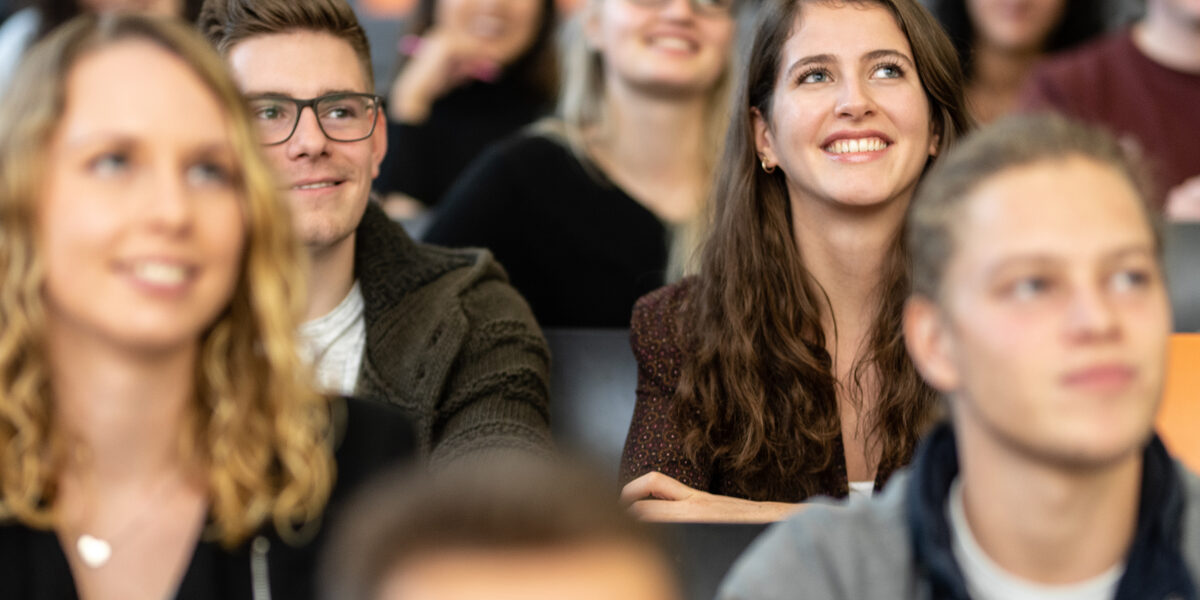 Nahaufnahme mehrerer Studierender in den Sitzreihen eines Hörsaals. Im Fokus eine Studentin und ein Student, die nebeneinander sitzen und geradeaus nach oben schauen.