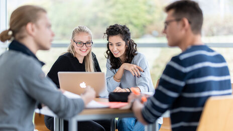 Photo of four students at a group table. The focus is on two female students sitting next to each other and looking at a laptop. Two other students are engrossed in a conversation.