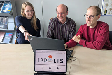 Three people are sitting in front of a laptop. The "IPPOLIS" logo can be seen on a monitor in front of them.